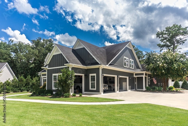 Fototapeta The side view of a large gray craftsman new construction house with a landscaped yard a three car garage and driveway