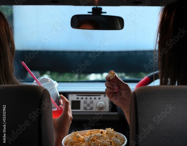 Fototapeta Two female friends with popcorn sitting in the car while watching a movie at drive in cinema. Selective focus. Entertainment, leisure activities, hobby concept