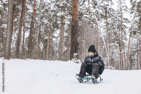 Fototapeta Happy teenager boy sledding and having fun outdoors. Joyful child playing in snow in winter forest. Laughing smiling kid walking in winter park in cold weather