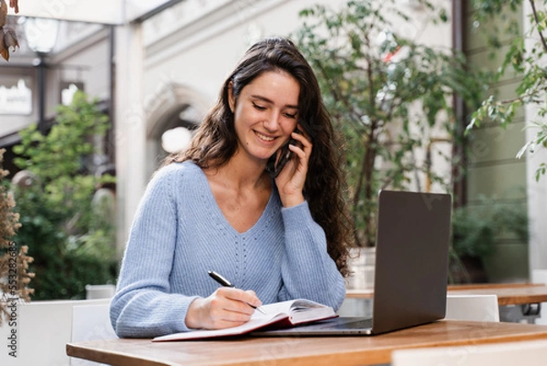 Fototapeta Happy girl is talking on the phone and chatting with colleagues remotely. Cheerful woman manager with laptop is making notes in notebook and talking with colleagues.