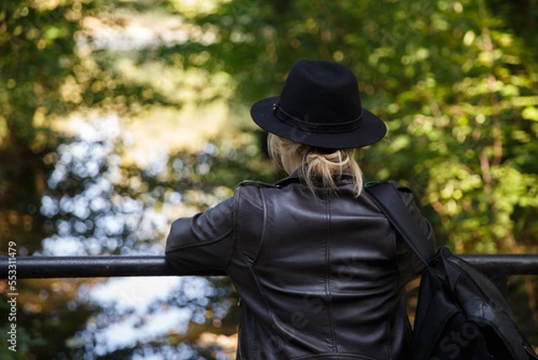 Fototapeta young Caucasian woman in a leather jacket, hat and with a backpack stands on the bridge and looks at the river. selective focus. High quality photo
