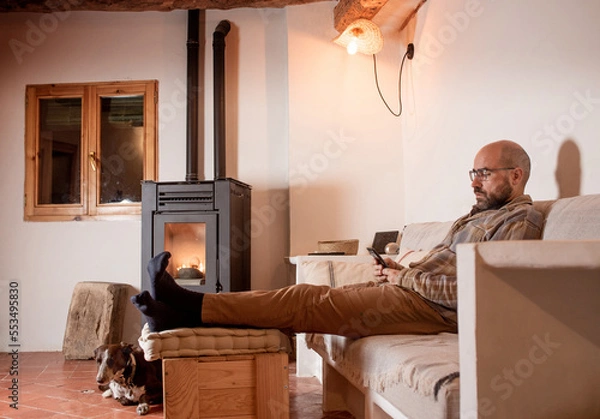 Fototapeta man in his forties looking at mobile phone, pellet cooker, dog resting on couch, petting dog. Rustic design dining room