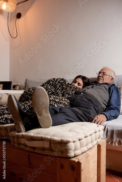 Fototapeta twenty-year-old granddaughter with her grandfather on the couch cuddling, snuggled up at home watching TV