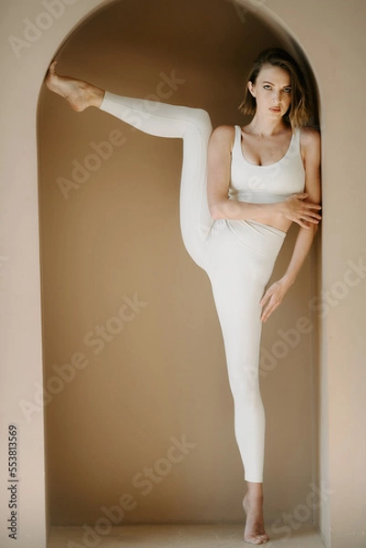 Fototapeta Front view of serious gymnastic female model, wearing in a white elastic body suit and stretching her leg in the unique pose while standing in arch in the wall of the beige wall in studio