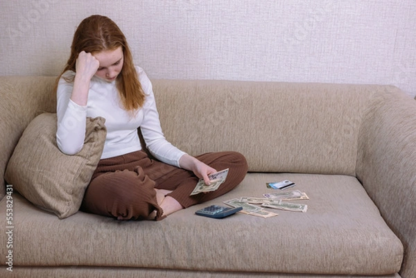 Fototapeta A young upset girl is counting the money of her budget sitting at home on the couch