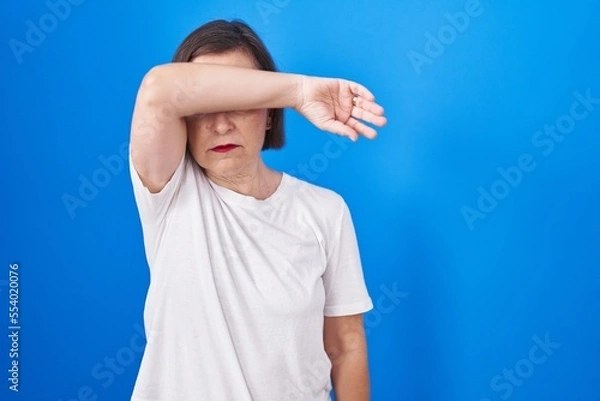 Fototapeta Middle age hispanic woman standing over blue background covering eyes with arm, looking serious and sad. sightless, hiding and rejection concept