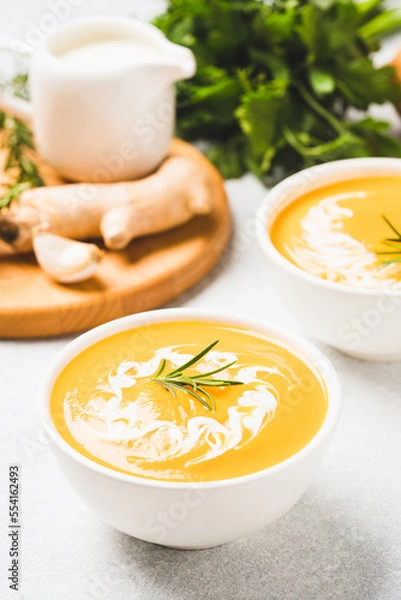 Fototapeta White bowl with thick butternut squash bisque garnished with heavy cream and rosemary on gray concrete table. Bowl of pumpkin cream soup with ginger. Vertical image, minimal style