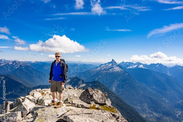 Fototapeta Adventurous athletic male hiker standing on top of a rugged mountain in the Pacific Northwest with jagged mountains in the background.