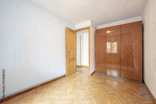 Fototapeta An empty room with a wardrobe with three-section glossy veneered doors with oak parquet floors placed in a herringbone pattern and hot water pipes on the skirting board