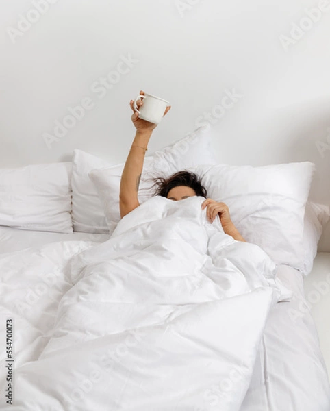 Obraz A woman shows her hand raised up while holding a cup of coffee behind a blanket in the bedroom. A young girl with a hand holding a cup sticking out from under the blanket. wake up with fun in the morn