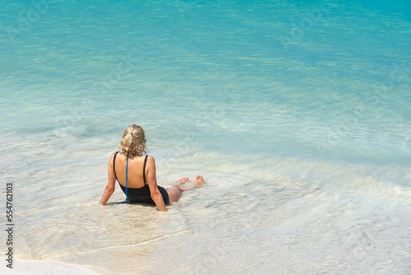 Fototapeta Rear view of young woman sitting on the sand by the ocean at the tropical beach enjoying water and ocean waves, copy space