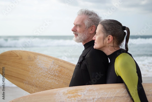 Fototapeta Side view of happy aged couple surfing together. Sporty man and woman in wetsuits standing on sea shore with surfboards, hugging, dreamily looking far. Sport activity, relations of aged people concept
