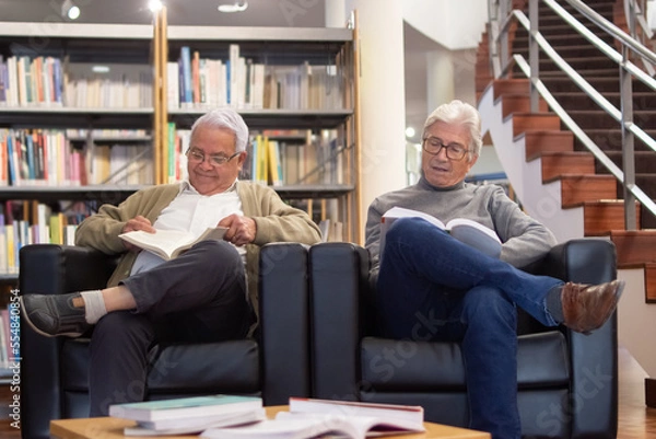 Fototapeta Positive aged men reading together. Two gray-haired students enjoying studying in university library finding information about technologies and discussing it. Studying of adult people concept