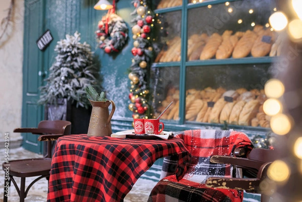 Obraz Table for two. Christmas bakery exterior with Christmas decorations and fresh bread in glass showcase. Cozy outdoor cafe on Christmas eve. Copy space. seasonal greetings concepts. Red cups of coffee.