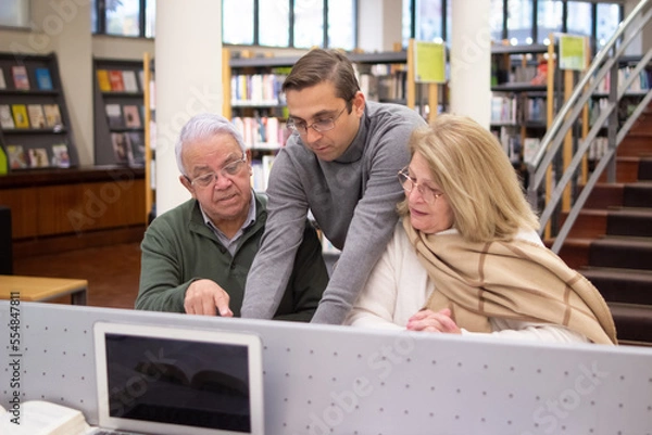 Fototapeta Portrait of aged couple getting knowledges. Serious man and woman sitting in library focused on reading books using computer listening to handsome instructor. Education for mature people concept