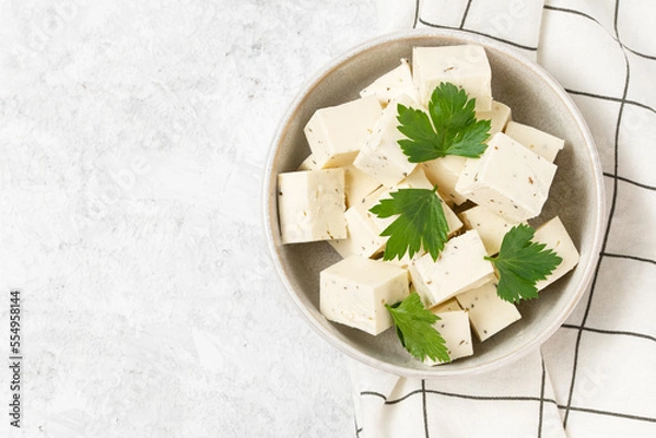 Fototapeta Tofu soy cheese or paneer or feta cheese cubes adding fresh parsley and celery in a ceramic bowl on a checkered napkin isolated top view