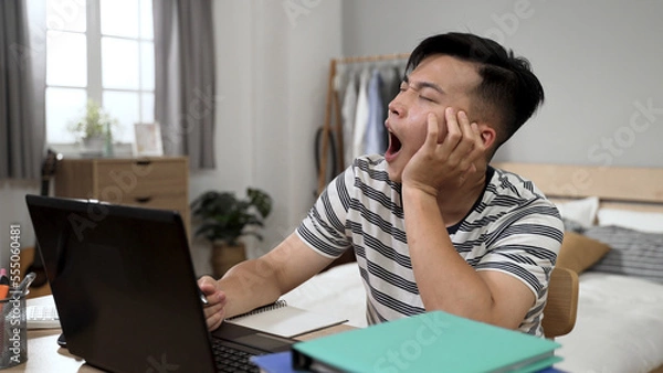 Obraz bored asian adult student propping face is playing with pen and looking away into distance with a yawn while learning from home on the computer in bedroom