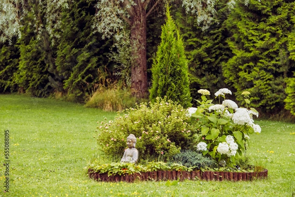 Fototapeta Meditating Buddha figurine sit inside small island of flowers and bush in flower bed, surrounded by natural wood roll palisade.