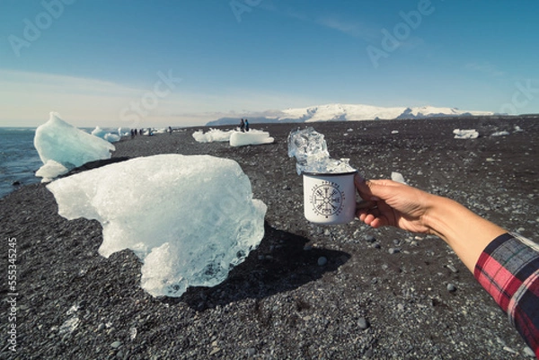 Fototapeta Close up tourist holding cup with ice on cold beach concept photo. First view hand photography with clear sky on background. High quality picture for wallpaper, travel blog, magazine, article