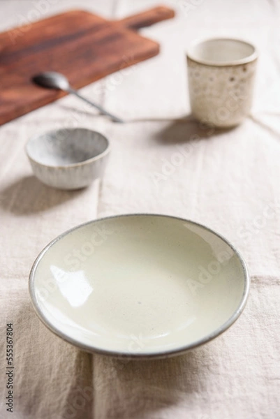 Fototapeta Empty pottery plate, sauce boat, cup, cutlery and cutting board on linen tablecloth. Table appointments. Selective focus