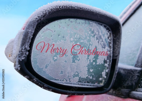 Obraz Red colour Merry Christmas handwriting or text on car side rear view mirror covered with frost against blurred snow and blue sky background, close up view.
