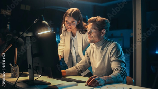 Fototapeta Software Developer Working on Desktop Computer in the Evening in Office. Young Female Project Manager Comes Over to the Desk to Discuss Updates to the New Online Application.