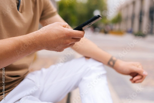 Fototapeta Close-up cropped shot of unrecognizable tattooed man sitting on bench using mobile phone looking to screen on city street. Unknow male in fashion clothes reading message on phone on summer day.