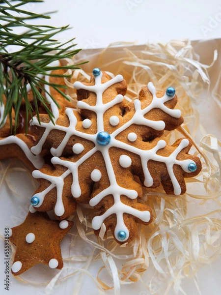 Fototapeta Christmas gingerbread cookies lie in a white box with straw near a pine branch. christmas cookies with icing. cookie in the form of a snowflake with blue dots. gingerbread lies on a white table.
