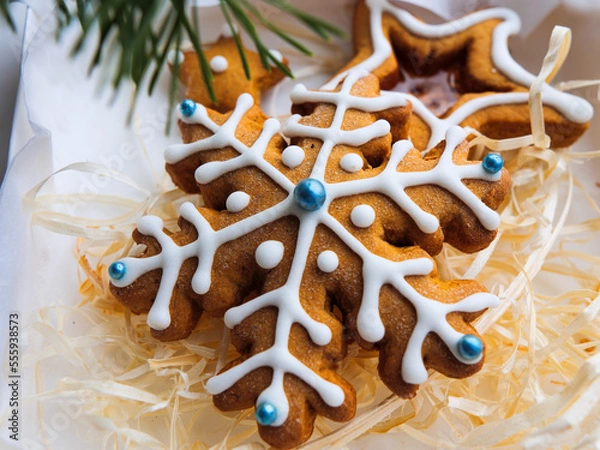 Obraz Christmas gingerbread cookies lie in a white box with straw near a pine branch. christmas cookies with icing. cookie in the form of a snowflake with blue dots. gingerbread lies on a white table.