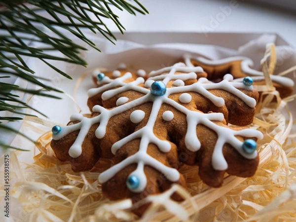 Obraz Christmas gingerbread cookies lie in a white box with straw near a pine branch. christmas cookies with icing. cookie in the form of a snowflake with blue dots. gingerbread lies on a white table.