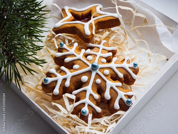 Obraz Christmas gingerbread cookies lie in a white box with straw near a pine branch. christmas cookies with icing. cookie in the form of a snowflake with blue dots. gingerbread lies on a white table.