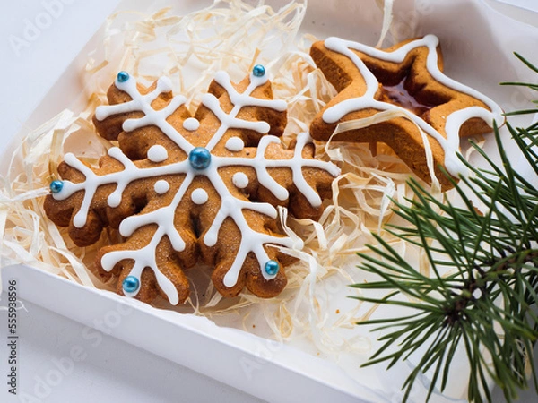 Obraz Christmas gingerbread cookies lie in a white box with straw near a pine branch. christmas cookies with icing. cookie in the form of a snowflake with blue dots. gingerbread lies on a white table.