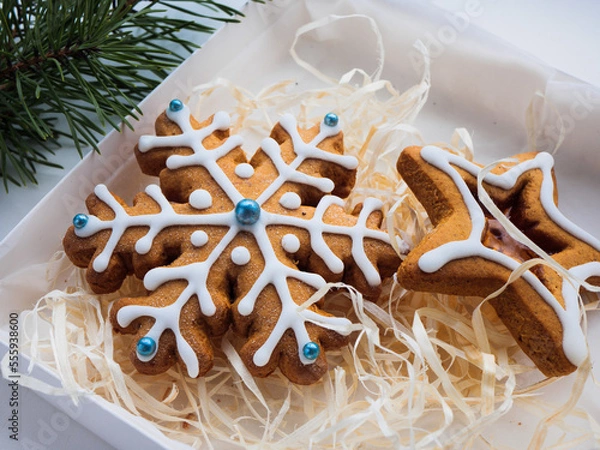 Obraz Christmas gingerbread cookies lie in a white box with straw near a pine branch. christmas cookies with icing. cookie in the form of a snowflake with blue dots. gingerbread lies on a white table.