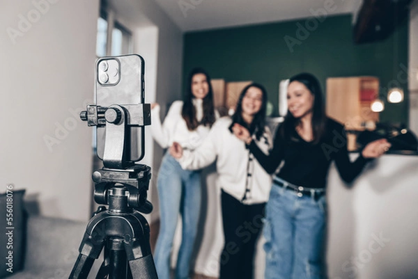 Obraz Close-up of a smartphone on a tripod filming 3 young white girls dancing, at home. Concept of making a living with videos on the Internet by being an influencer. Popularity and celebrity online