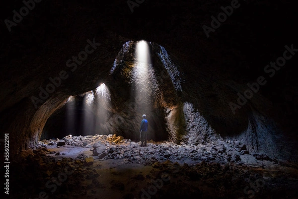 Fototapeta Adventurous athletic male standing in a lava tube looking at the sunlight shinning down into the cave through holes in the ceiling.
