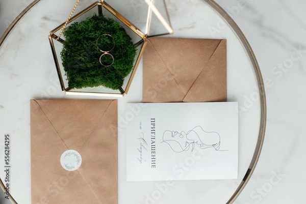 Fototapeta The concept of love and relationships. Wedding invitation. Gold rings in a gold casket on a marble table. Craft envelopes with invitations. Close-up.