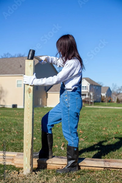 Fototapeta woman diy enthusiast wearing overalls installing a wooden fence on backyard. She is hammering a 4x4 post on the ground
