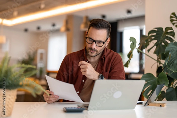 Fototapeta Serious millennial man using laptop sitting at the table in a home office, looking at the paper, communicating online, writing emails, distantly working or studying on computer at home.