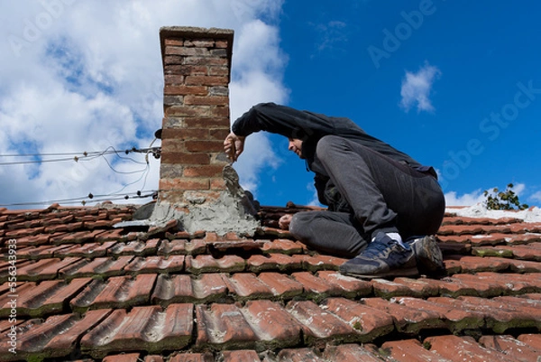 Obraz A skilled craftsman on the roof of an old house, using a trowel to repair the chimney. The man sits focused on the task at hand. Sky in background
