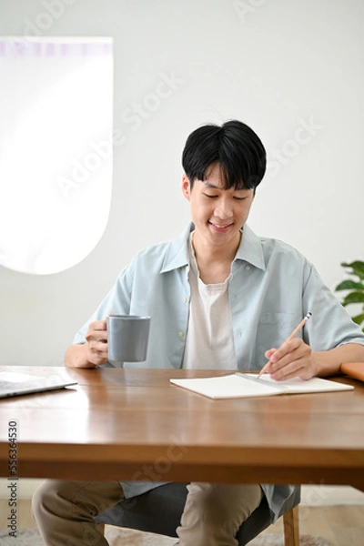 Fototapeta Handsome young Asian male college student sipping coffee while focusing on his homework