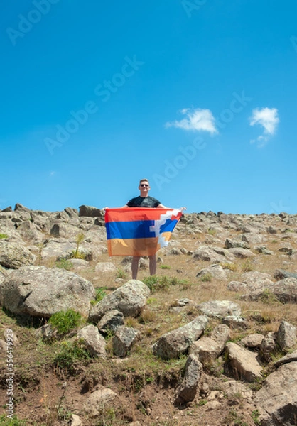 Fototapeta a young handsome man holds in his hands the flag of the unrecognized republic of Nagorno-Karabakh or Artsakh, standing in a large steppe with piled large stones. Support for Artsakh