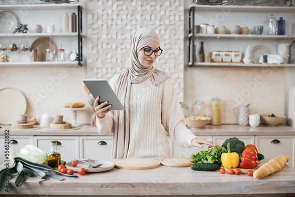 Fototapeta Happy muslim woman in hijab cooking healthy food pointing on vegetables lying on counter while reading recipe on digital tablet in modern kitchen at home. Preparing vegetable salad.