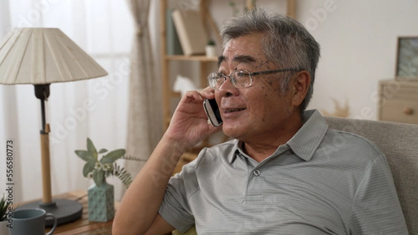 Obraz Smiling asian old man talking on smartphone on sofa. Mature male in online conversation using mobile phone while laughing. cheerful elderly guy in glasses having fun during funny conversation call