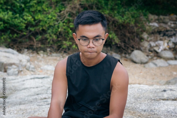 Fototapeta Pensive Filipino man with eyeglasses looking nervous sitting alone on rocks. Worried male adult with thoughtful look, solitude concepts