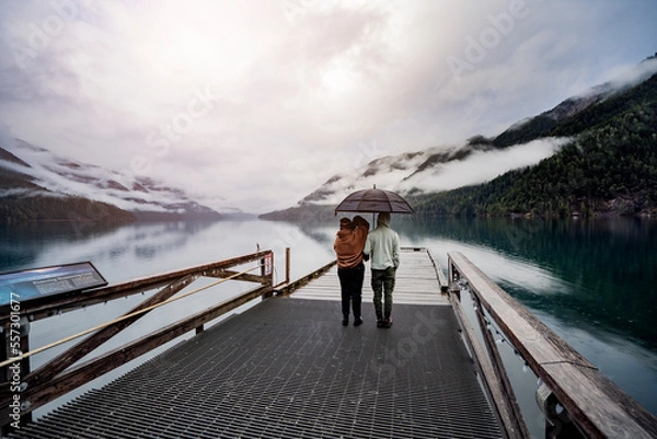 Fototapeta 
A young sympathetic couple stands with an umbrella in their hands against the backdrop of a beautiful lake and mountains.