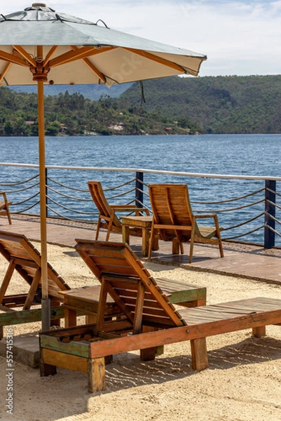 Fototapeta Empty wooden lounge chairs on the sand under green umbrella. In the background, a huge lake. Vertical