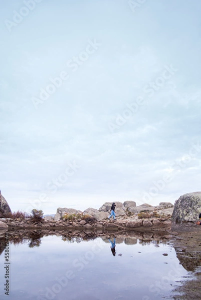 Obraz latin woman taking a picture of herself on some rocks that cross a lagoon, where you can see the reflection of the sky and the woman.