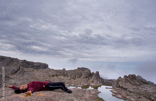 Fototapeta latin woman lying on a rock while smiling, with a lagoon and a valley of rocks in the background.