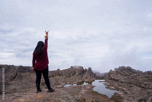 Obraz latin woman with her back turned and her arm raised with the sign of the metal, standing on the edge of a cliff with a lagoon in the background.