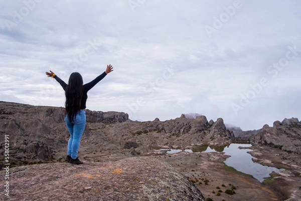 Obraz Latin woman with her back turned and arms raised, standing on the edge of a cliff with a lagoon in the background.